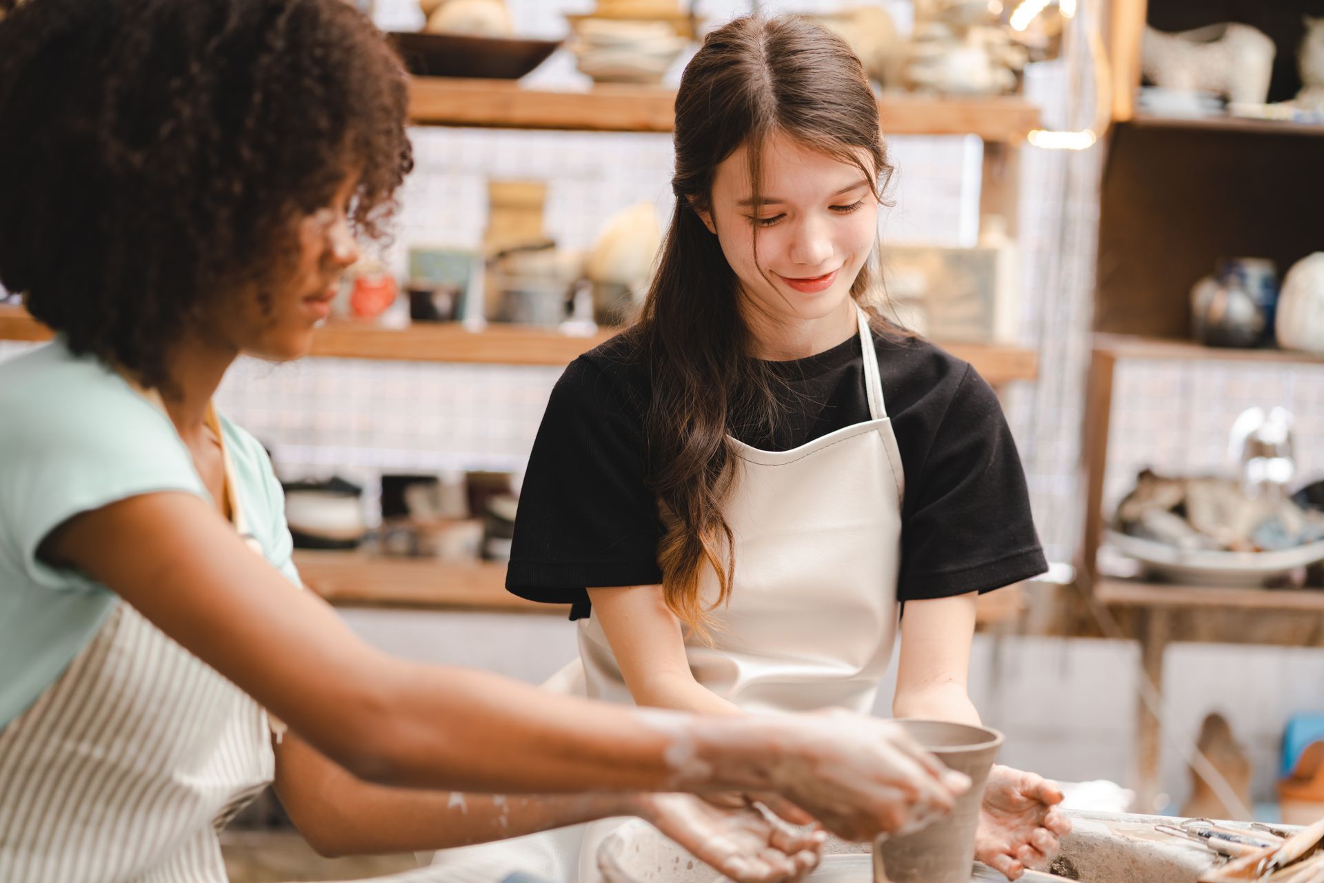 Two people in a pottery studio. One helps the other shape a clay pot on a wheel.