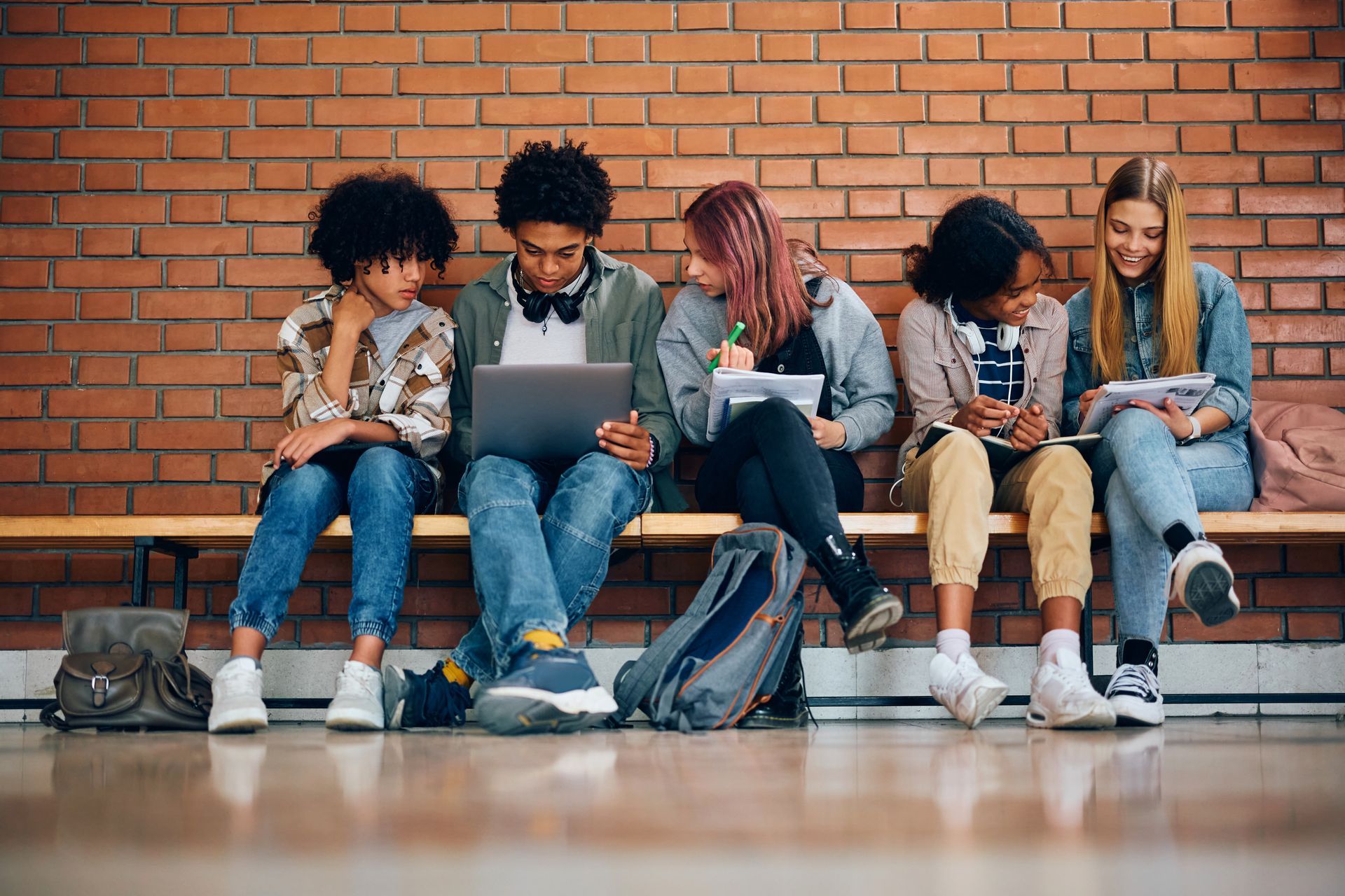 Students on a bench in front of a brick wall, some looking at laptop and other devices.