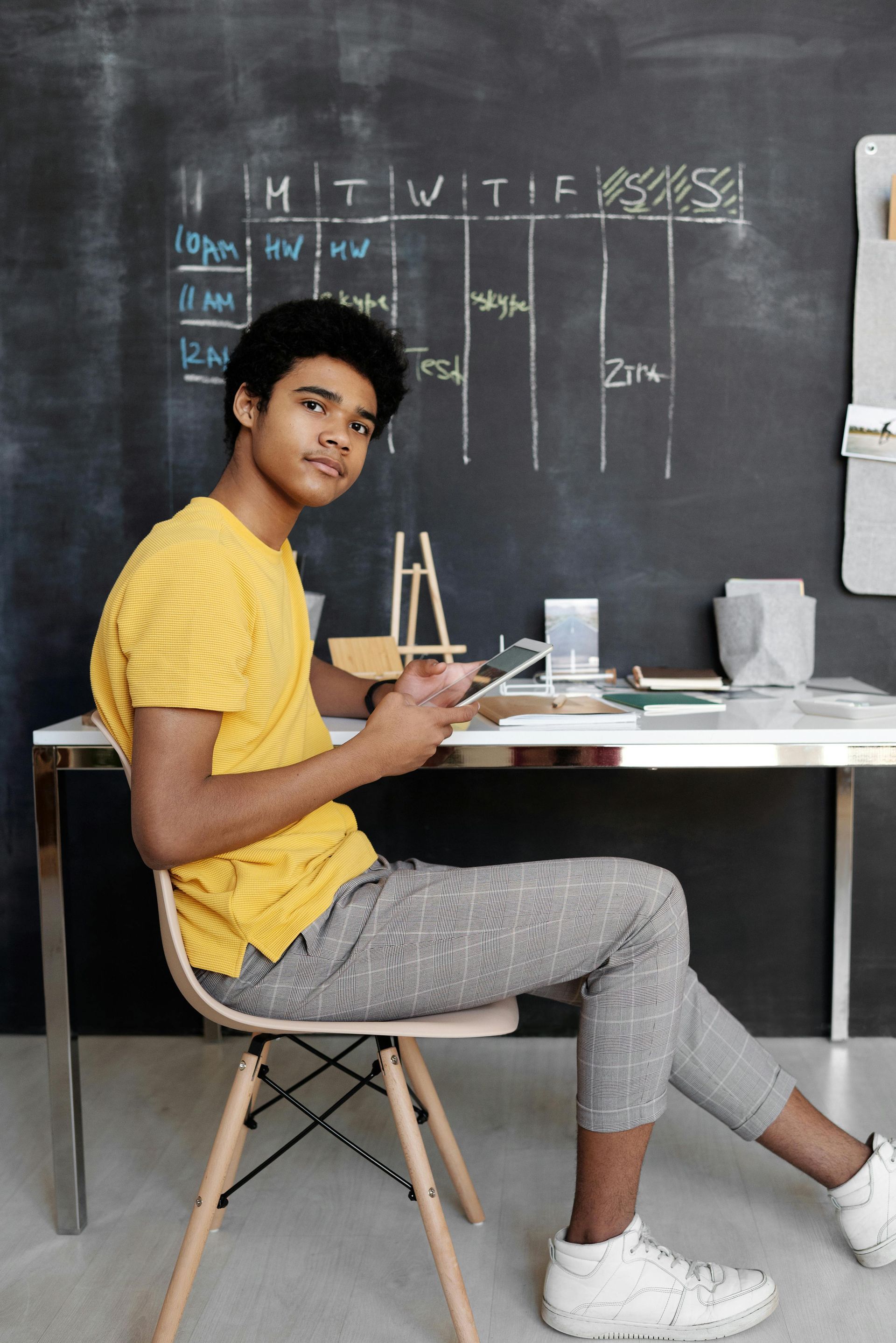 Young person seated at desk, holding tablet. Yellow shirt, patterned pants, against a chalkboard.