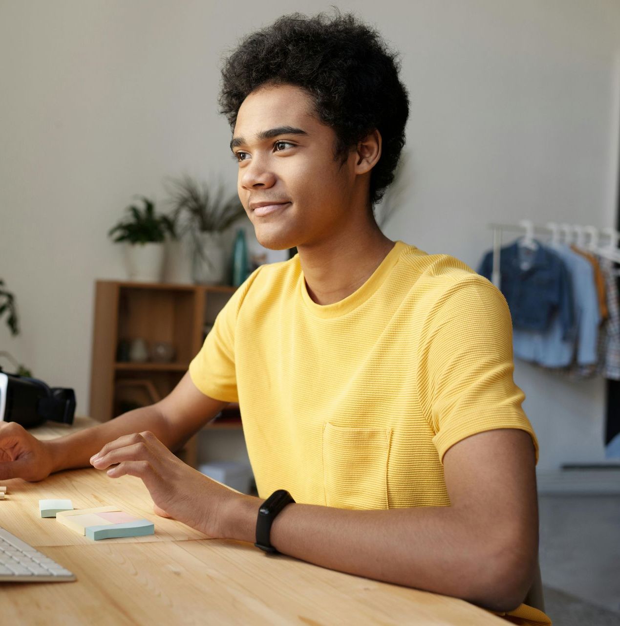 Person with curly hair wearing a yellow shirt, smiling while working on a computer at a wooden desk.