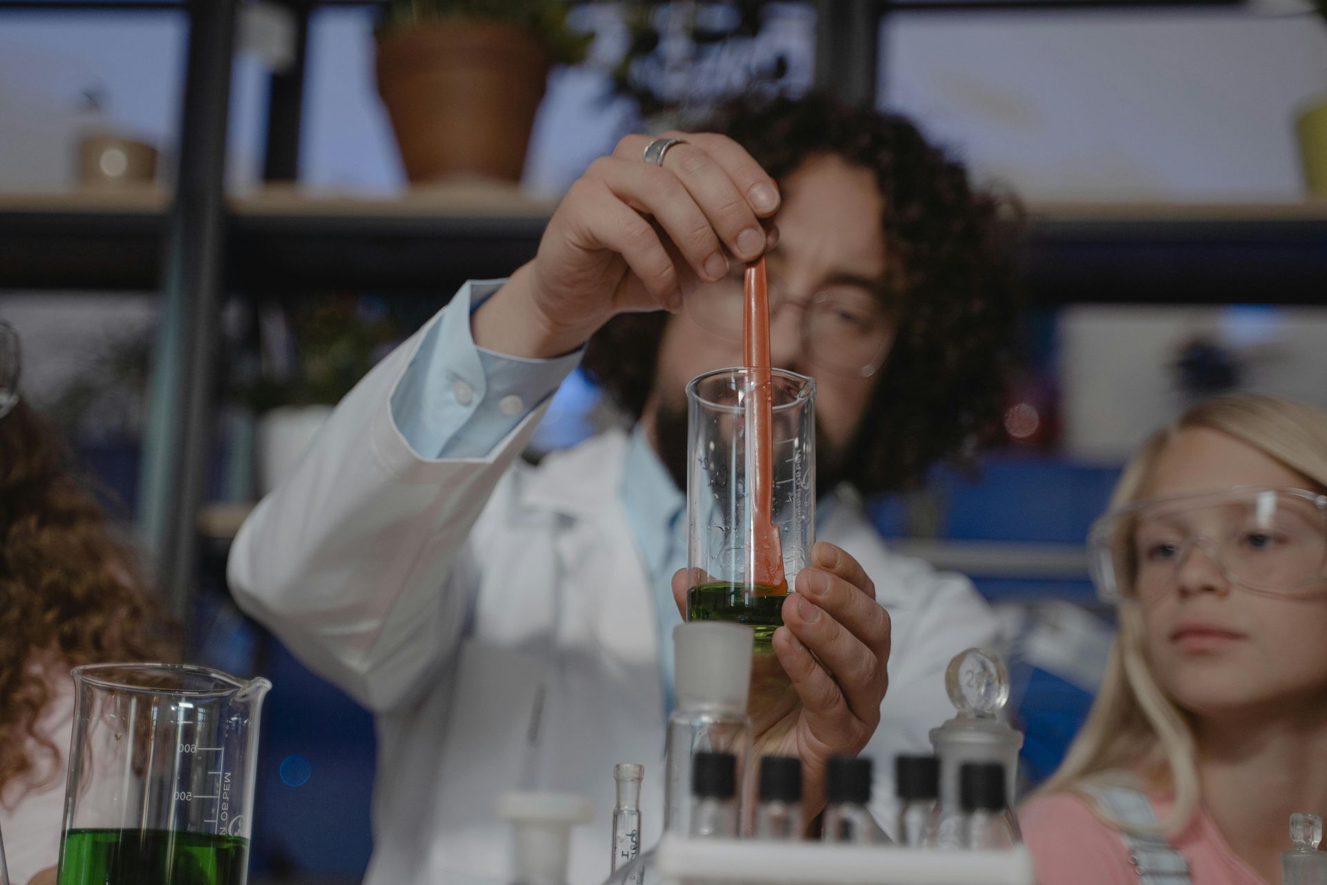 Scientist in lab coat stirring liquids in glass tubes; two children watch.
