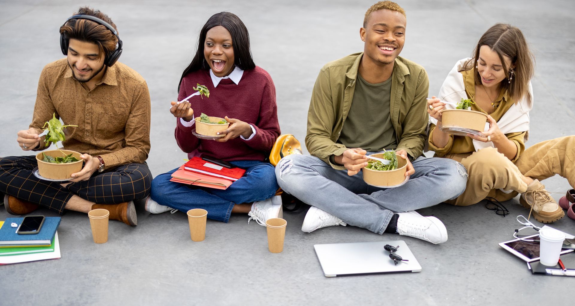 Four people seated outdoors, eating salad from containers, smiling.