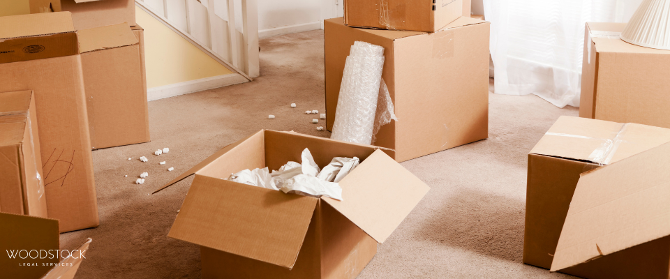 Cardboard moving boxes scattered around a carpeted room, some open with packing paper and bubble wrap visible, suggesting a home move or packing process.