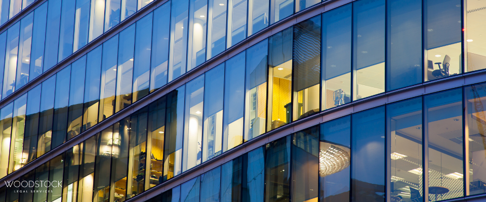 A modern glass office building with blue-tinted reflective windows and illuminated interiors. The perspective emphasizes the building’s diagonal architectural lines and sleek corporate design. Small interior details, including desks, lights, and furnishings, are visible through the windows. The lower-left corner contains the text “WOODSTOCK LEGAL SERVICES.”
