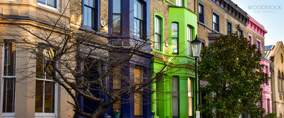 A vibrant row of traditional Victorian terraced houses, each painted in a different bold colour, including beige, deep purple, bright lime green, and pink. The architectural details feature classic bay windows and decorative masonry, typical of London neighbourhoods like Notting Hill. A leafless tree stands in the foreground, and a vintage-style street lamp is positioned in front of the greenhouse. The logo for