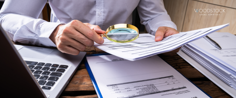 Business professional examining financial documents with a magnifying glass at a desk, alongside a laptop and paperwork, with Woodstock Legal Services logo in the background