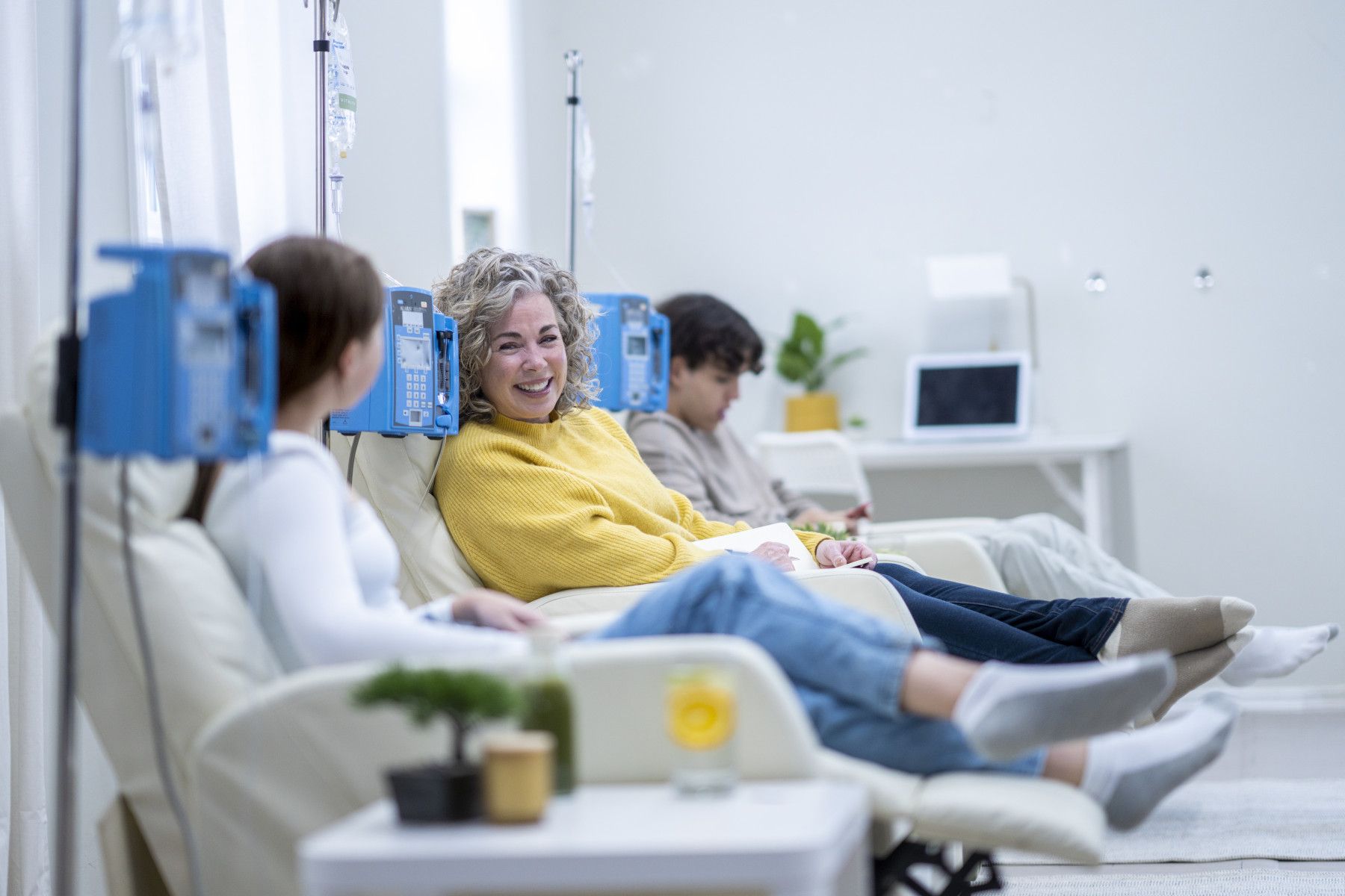 People seated in a bright medical waiting room, with an IV stand and wall-mounted monitors nearby