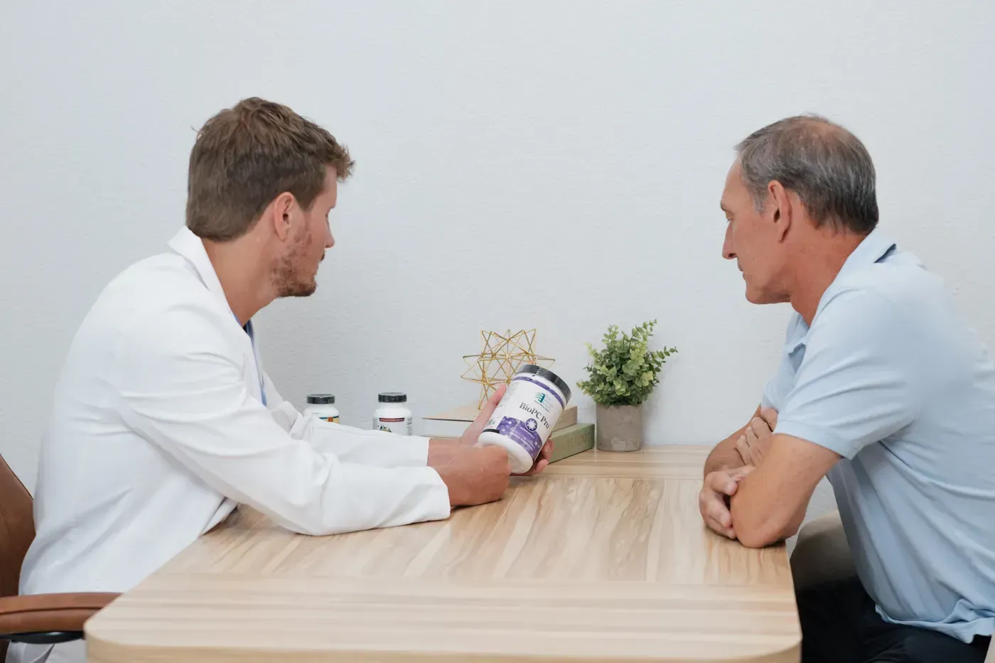 Two people in white coats discuss a jar of pills across a table in a clinic office.