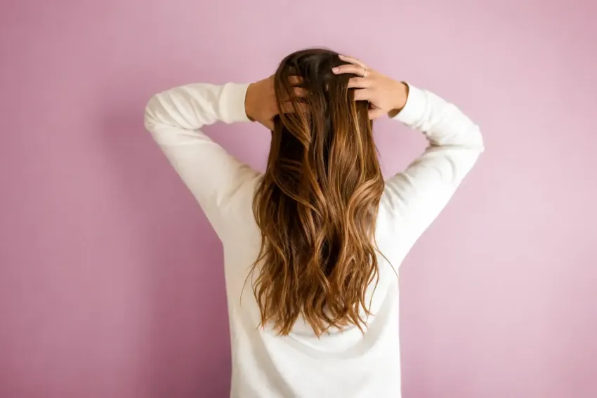 Person with long brown hair standing with hands on head against a pink wall