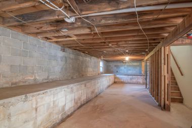An unfinished, empty basement featuring exposed wooden ceiling joists, concrete block walls, and a staircase.