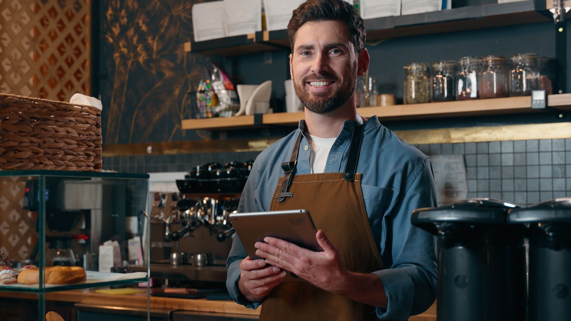 Smiling barista holding a clipboard behind a coffee shop counter with espresso machines and shelves.