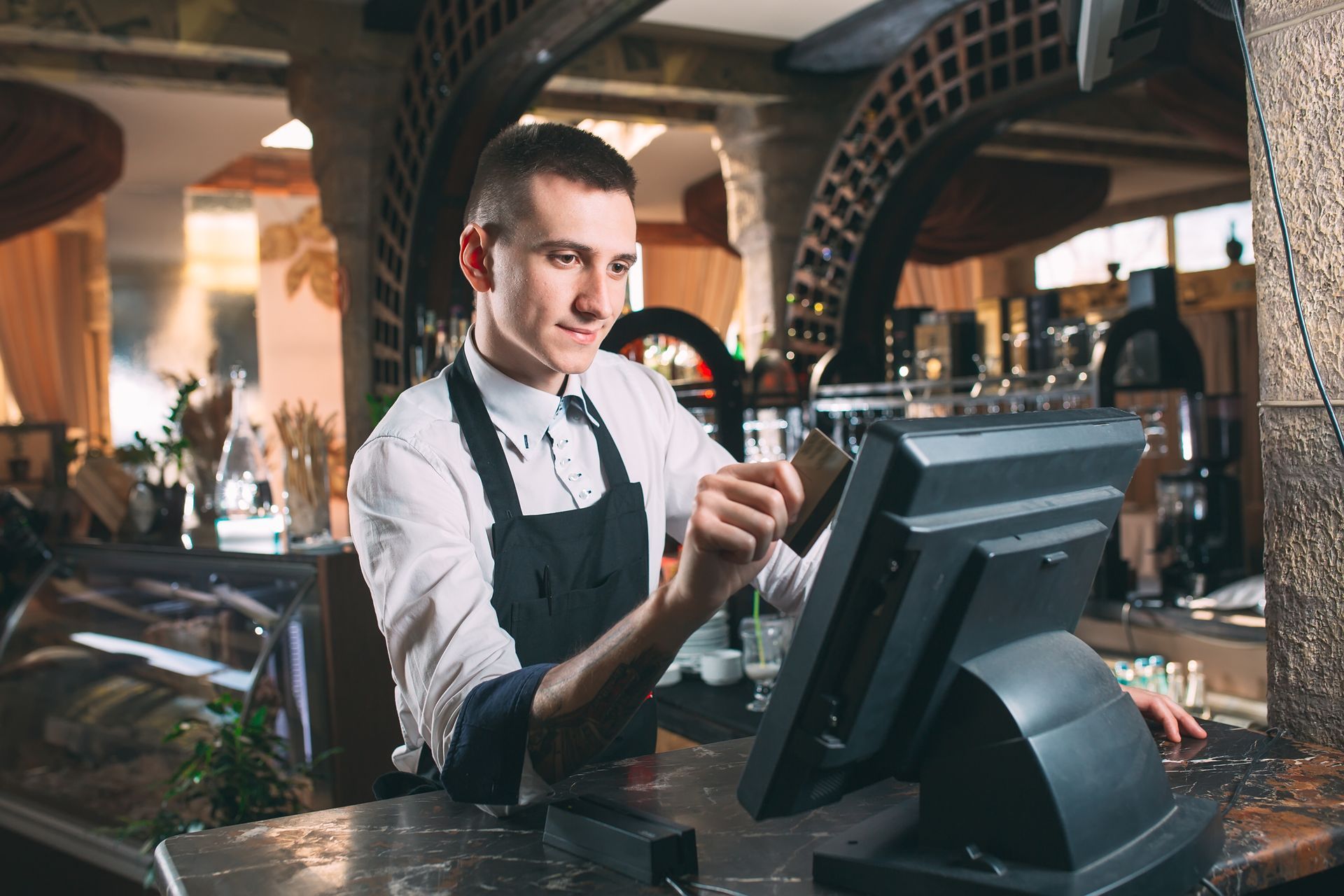Bartender at a bar tapping a card on a POS terminal with bottles and glasses in the background