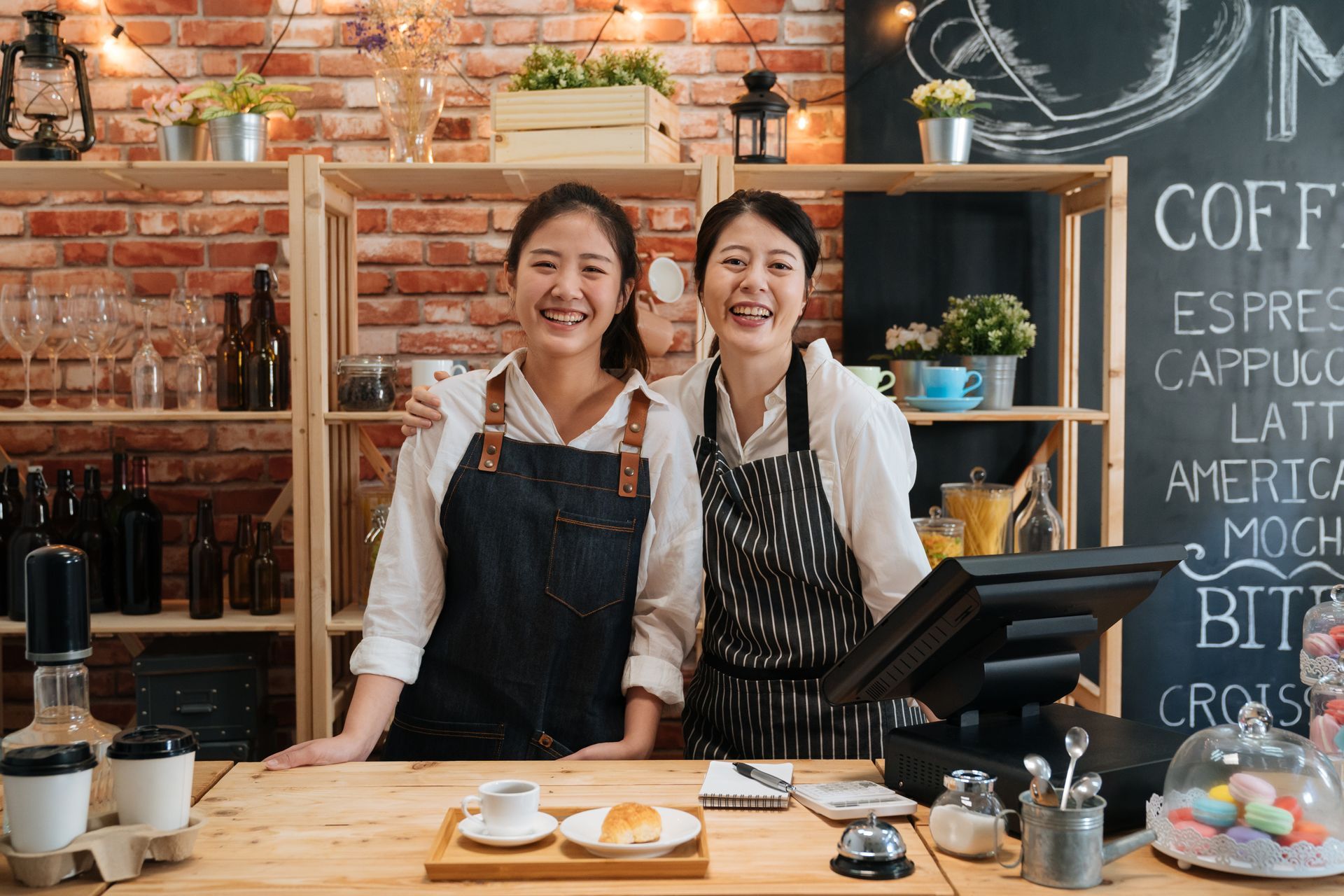 Two smiling baristas behind a coffee shop counter, with chalkboard menu and espresso equipment.