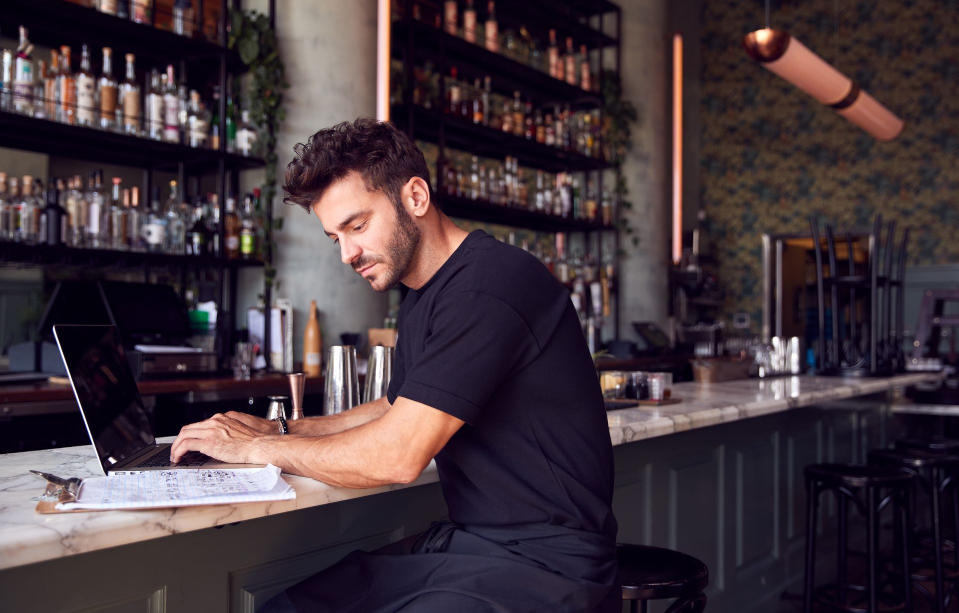 Man typing on a laptop at a bar counter with bottles and glasses on shelves behind him