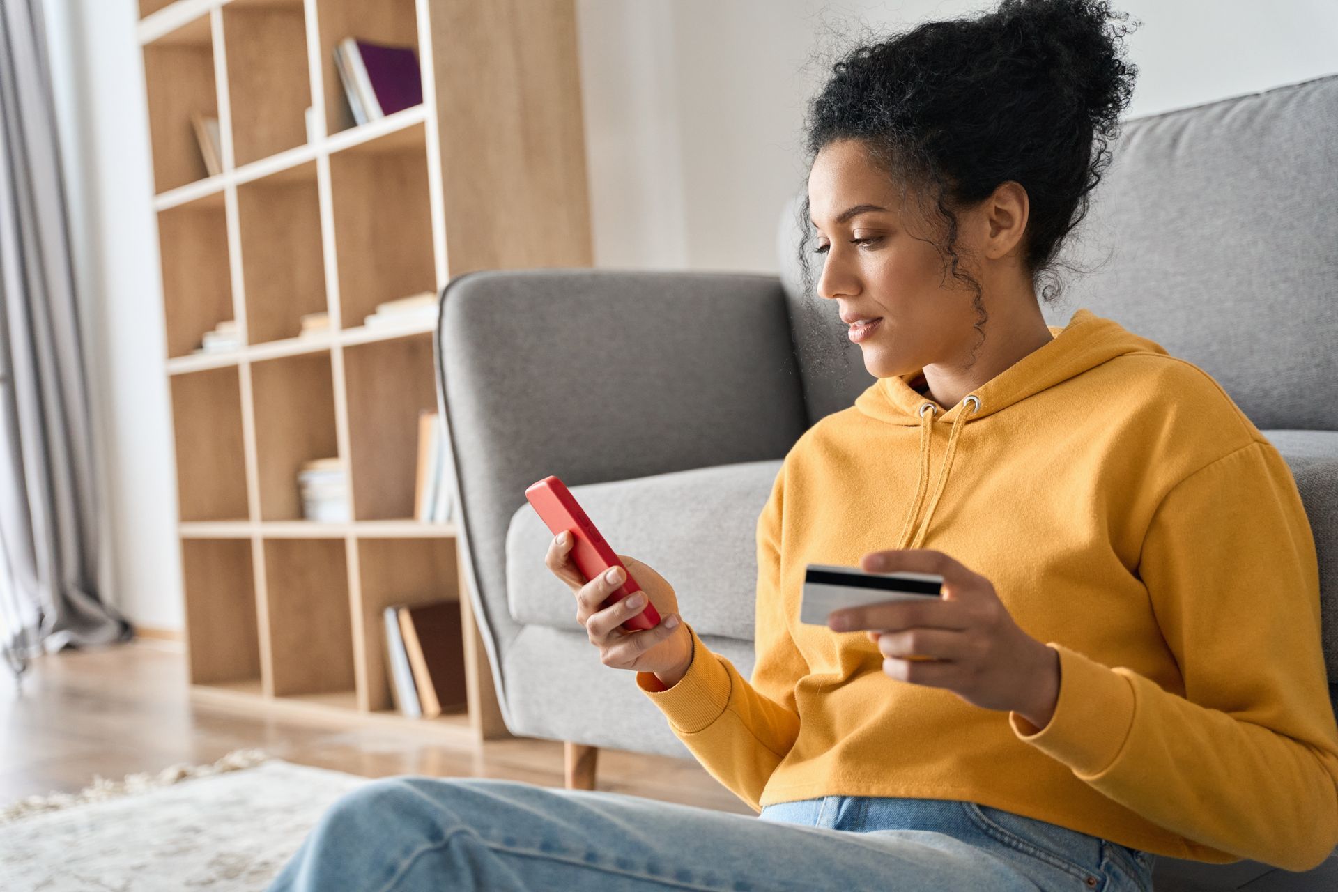 Woman on a couch holding a red phone and black card, looking at her screen in a bright living room.