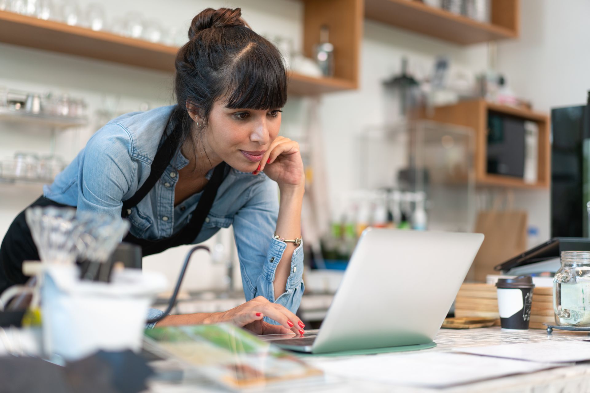 Woman leaning over a laptop in a bright kitchen, focused on work with papers and coffee nearby
