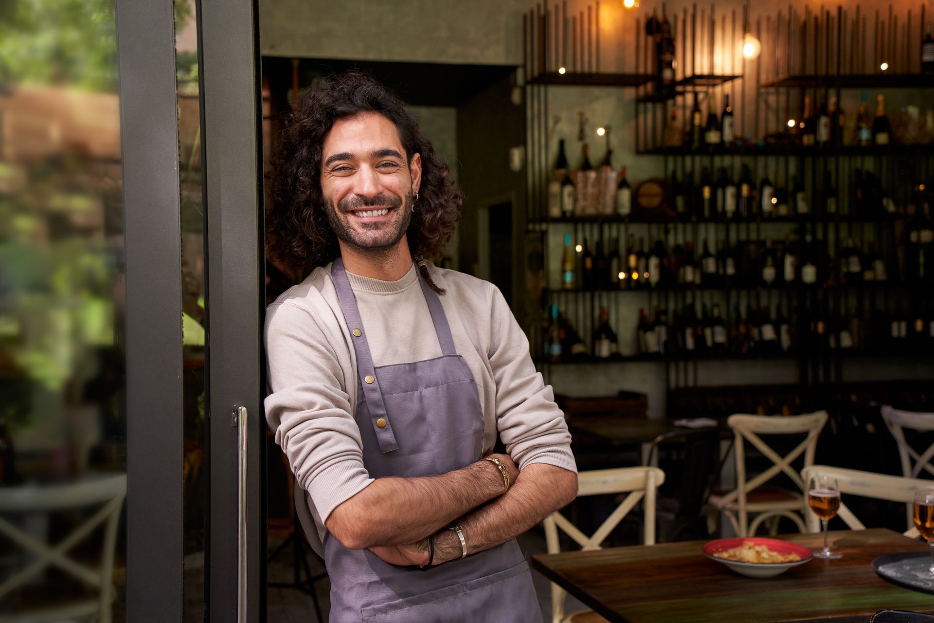 Smiling chef in an apron standing in a cozy restaurant with tables and bottles behind him