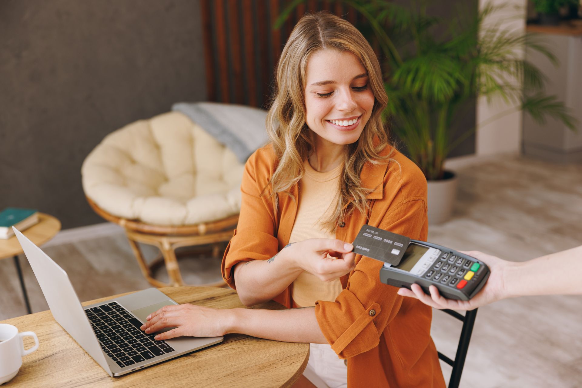 Woman smiling and tapping a card reader beside a laptop in a cozy room