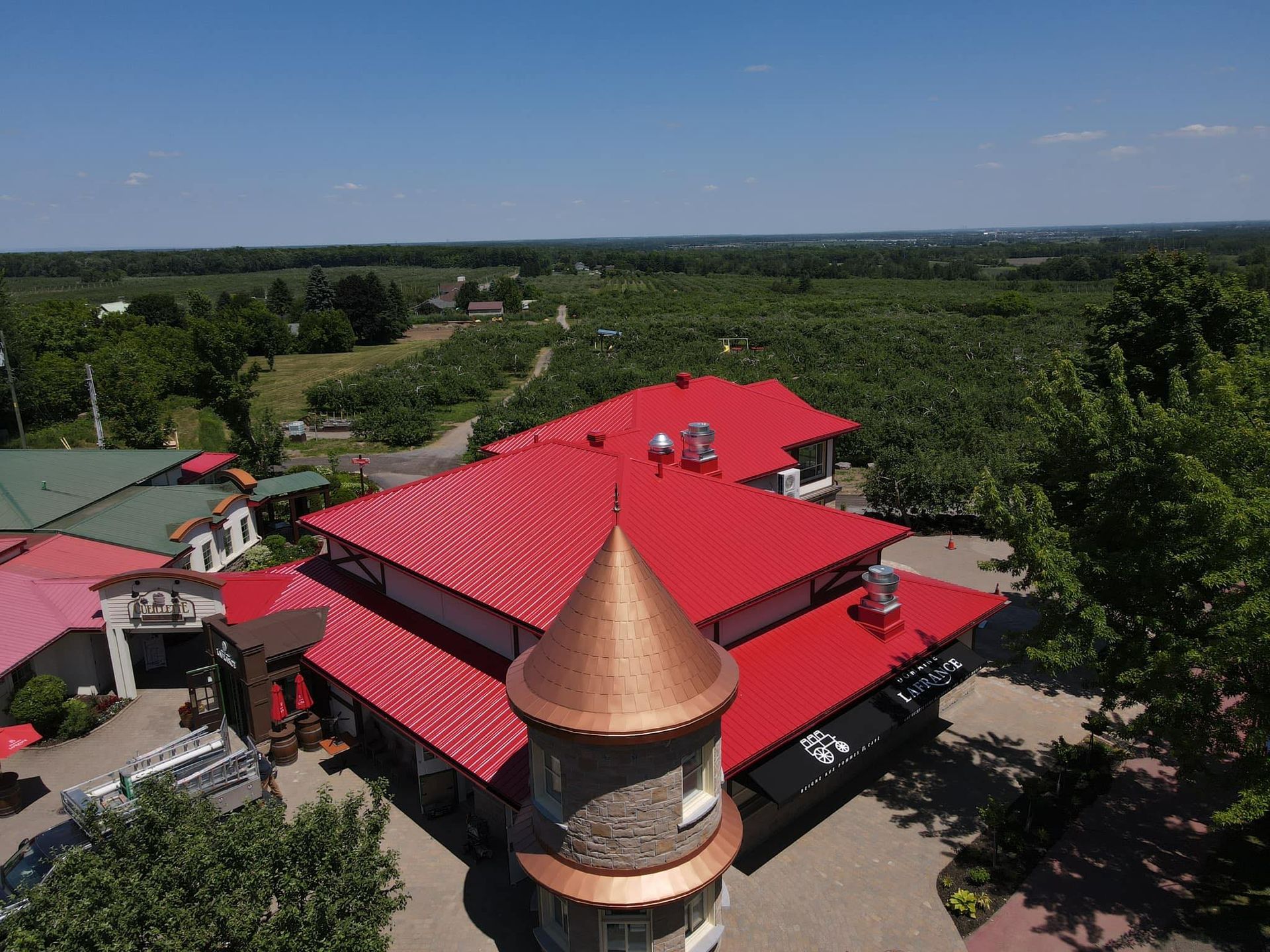 Vue aérienne d'un bâtiment au toit rouge avec une tourelle, entouré d'arbres et de champs.
