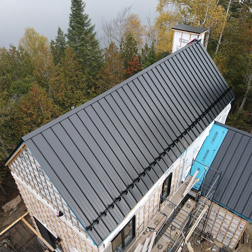 Vue aérienne d'une maison en pierre avec un toit en métal sombre en construction, entourée d'arbres d'automne.