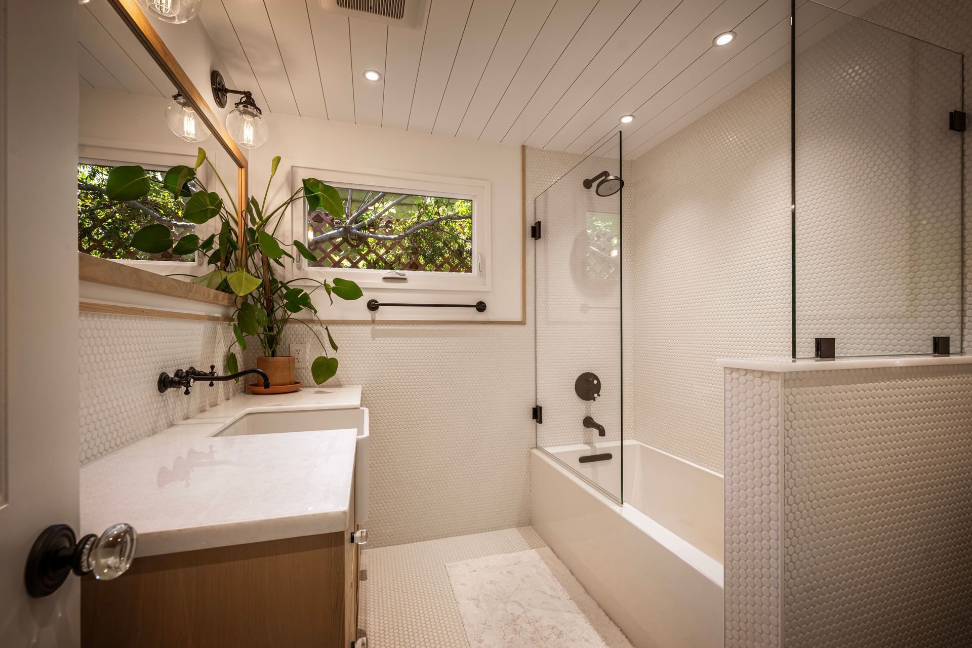 A bright bathroom with white penny tiles, a wood vanity, a glass-enclosed bathtub, and a potted plant by the window.