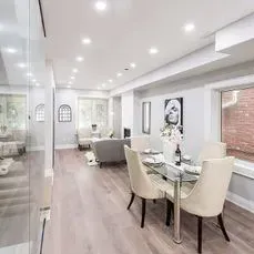 Dining area with glass table, four cream chairs, light wood floor, and a living room in the background.
