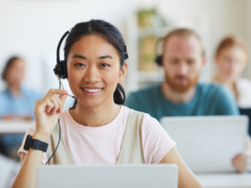 Smiling customer support agent wearing a headset in front of a laptop, with coworkers blurred in the background