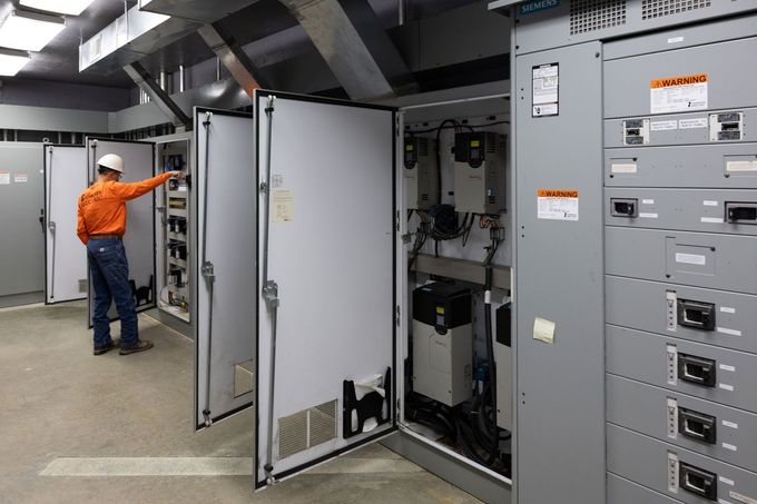 A man in an orange shirt is working on a electrical panel