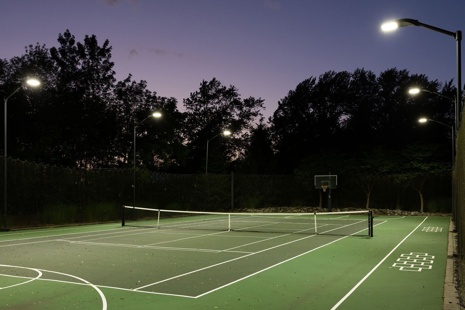 A tennis court is lit up at night with trees in the background