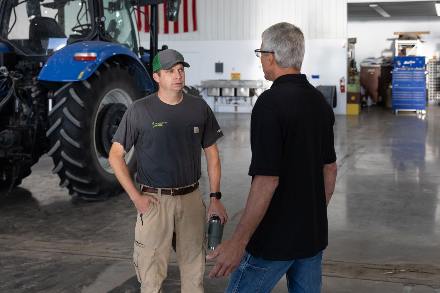 Two men are talking in a garage in front of a tractor.