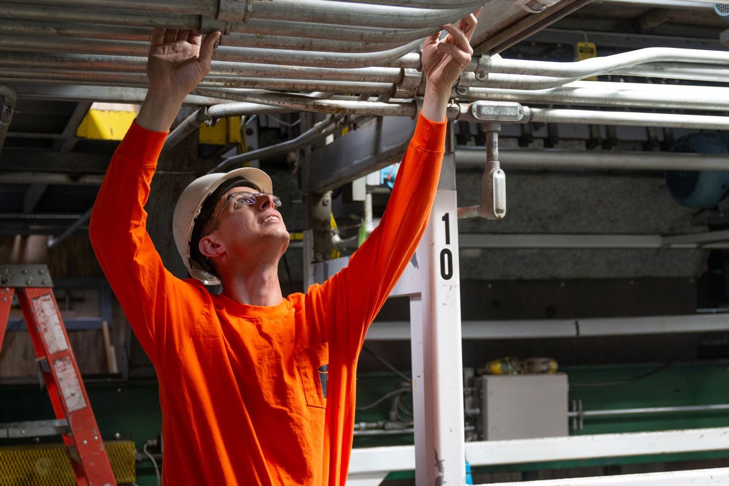 A man in an orange shirt and hard hat is working on a pipe.