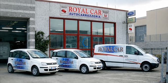Furgoni bianchi della Royal Car parcheggiati davanti a un'officina meccanica con vetrina in una via della città.