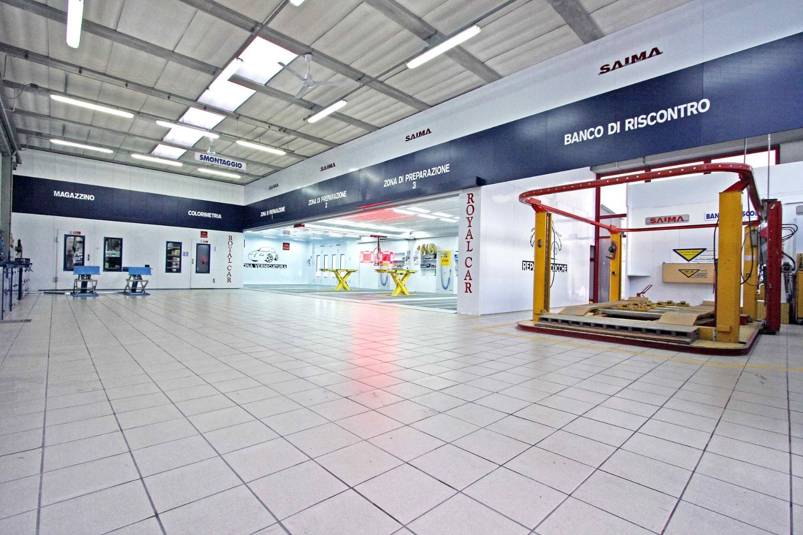 Ampia officina meccanica al coperto con pavimento piastrellato, veicoli sollevati su ponte e segnaletica in spagnolo lungo le pareti.