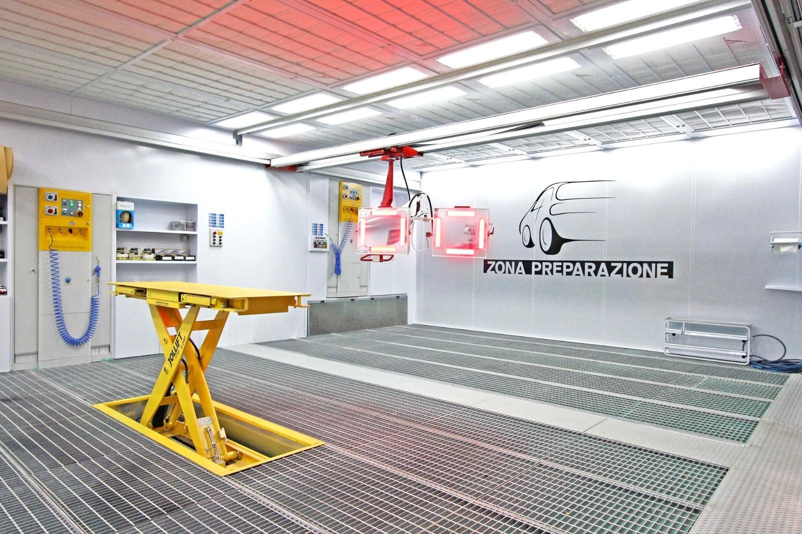 Interno di un'officina industriale con piattaforma elevatrice gialla, attrezzature aeree rosse e grigliato metallico sul pavimento.