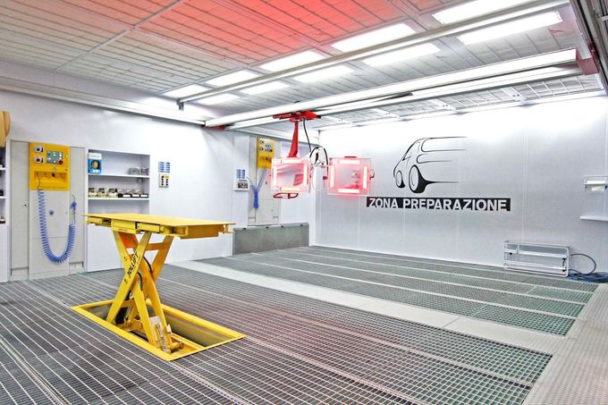 Cabina di verniciatura industriale luminosa con elevatore giallo, griglie metalliche e attrezzature aeree rosse.