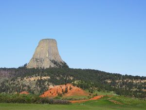 A flat-topped rock mesa rises above green trees and red soil under a clear blue sky, Devils Tower National Monument, Wyoming