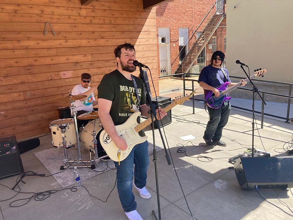 Three musicians perform outdoors on a patio, with guitars, drums, and microphones.