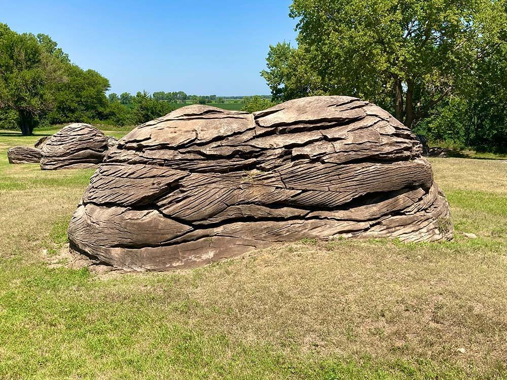 Large layered rock formation in a grassy field under a blue sky, with trees in the background, Rock City, Minneapolis, Kansas