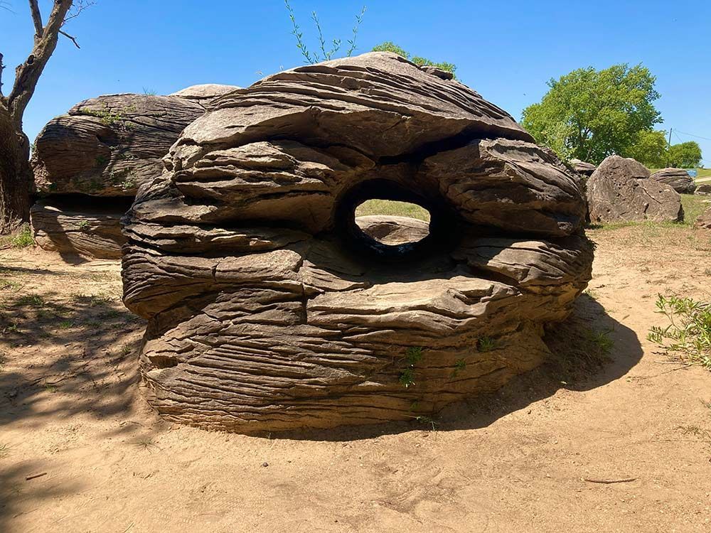Stacked rock formation with a circular hole in a sandy, sunlit landscape under a clear blue sky, Rock City, Minneapolis, Kansas