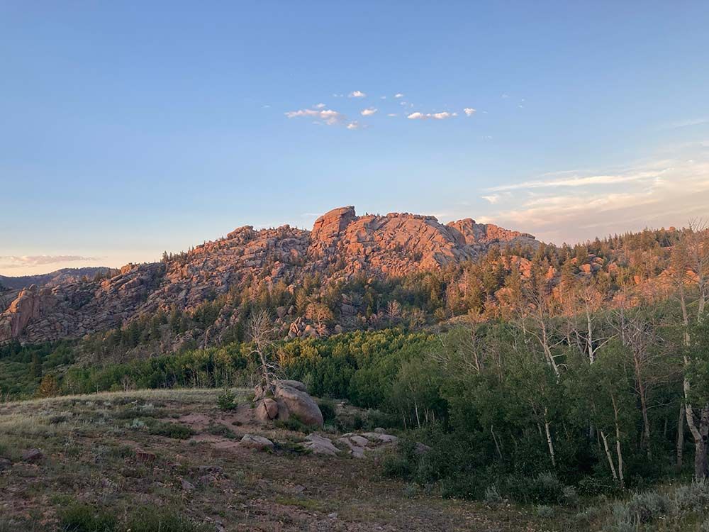 Sunlit mountain ridge at sunset beneath a clear blue sky, with forested hills in the foreground, Vedauwoo, Wyoming