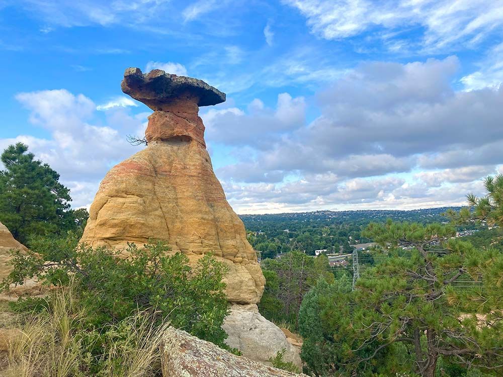 Tall mushroom-shaped rock formation under a blue sky with scattered clouds, surrounded by green trees, Palmer Park, Colorado Springs, Colorado