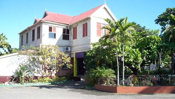 Two-story white house with red roof and shutters, surrounded by tropical trees and plants in bright sunlight