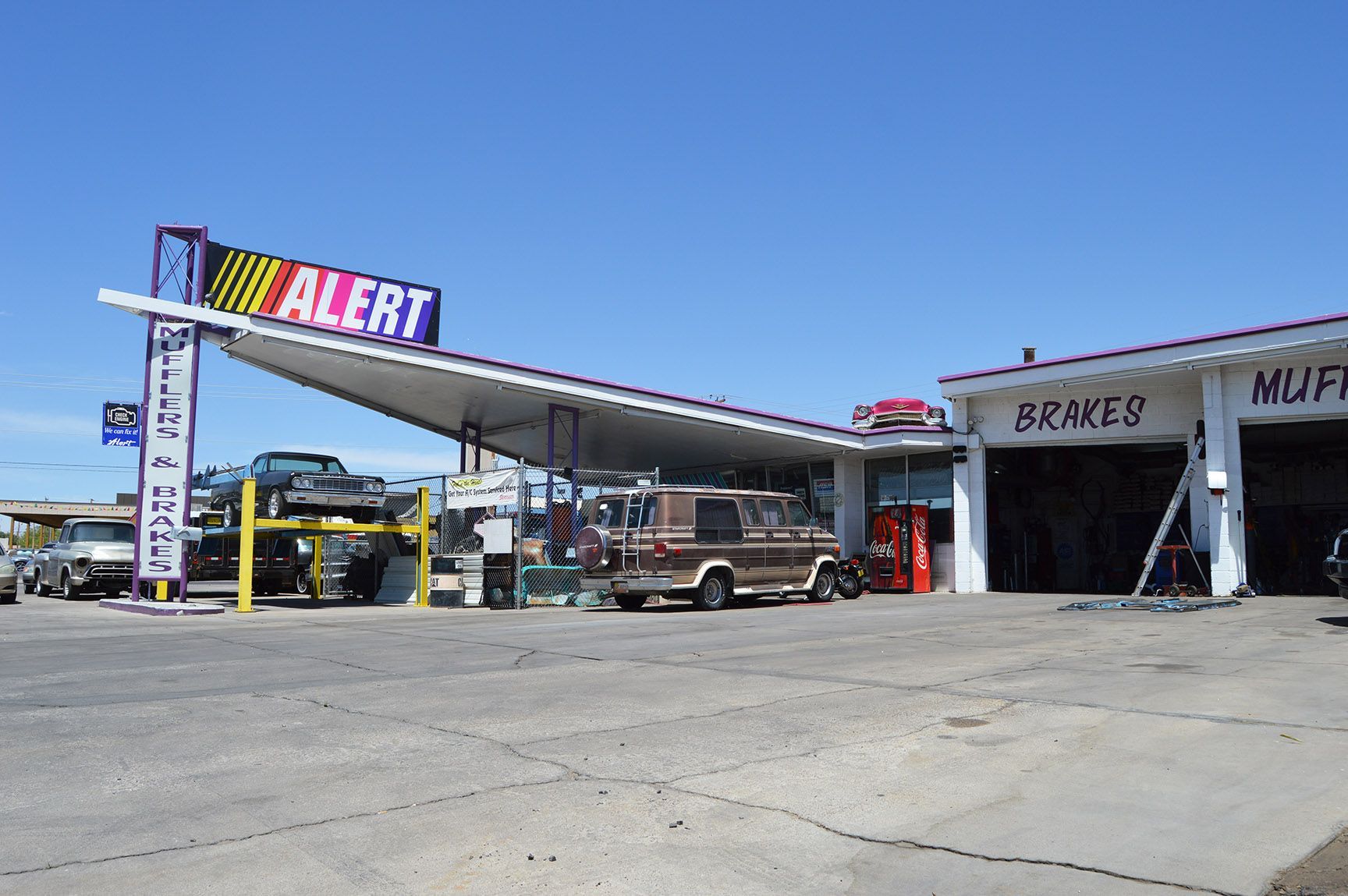 Outdoor auto repair shop with car lift, trucks, and a shaded canopy under a clear blue sky