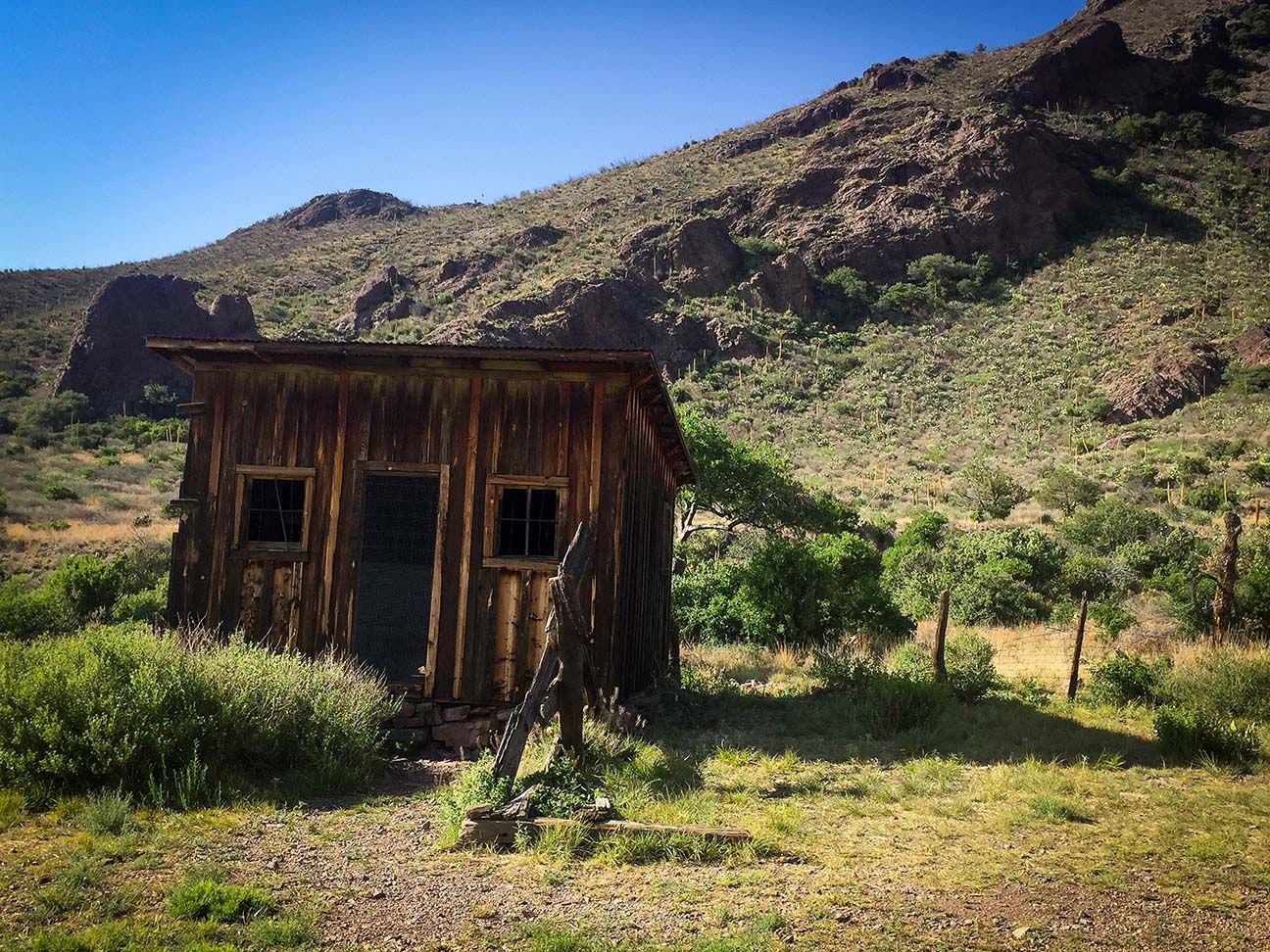 Small weathered wooden shack in a sunny, grassy desert valley with rocky hills behind it.