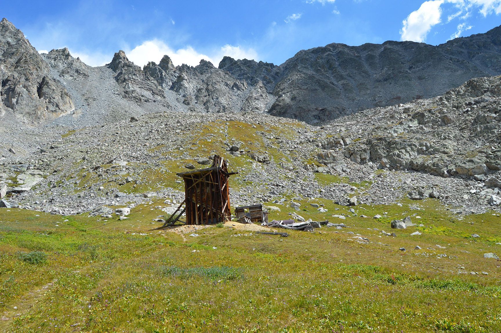 Grassy alpine meadow with a small wooden hut beneath rocky mountain cliffs and a blue sky.Mayflower Gulch, Colorado, Gold Crest Mine