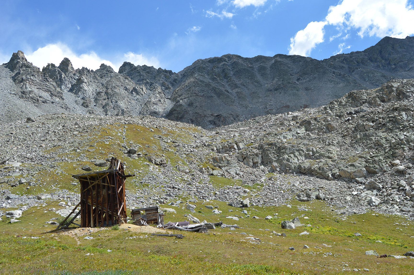Rocky mountain slope under blue sky, with a small rusted wooden structure in the foreground. Mayflower Gulch, Colorado, Gold Crest Mine