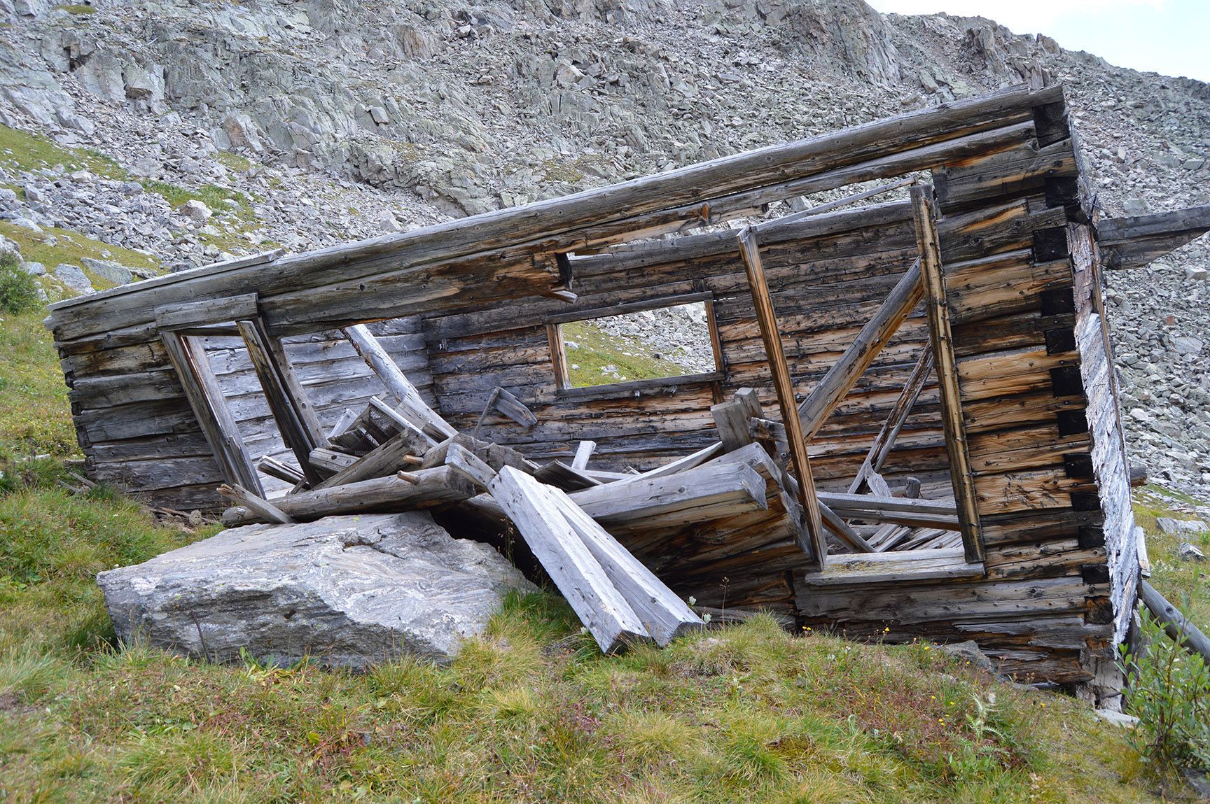 Collapsed rustic wooden shed on a grassy mountainside, with stone rubble and a rocky slope behind it, Mayflower Gulch, Colorado