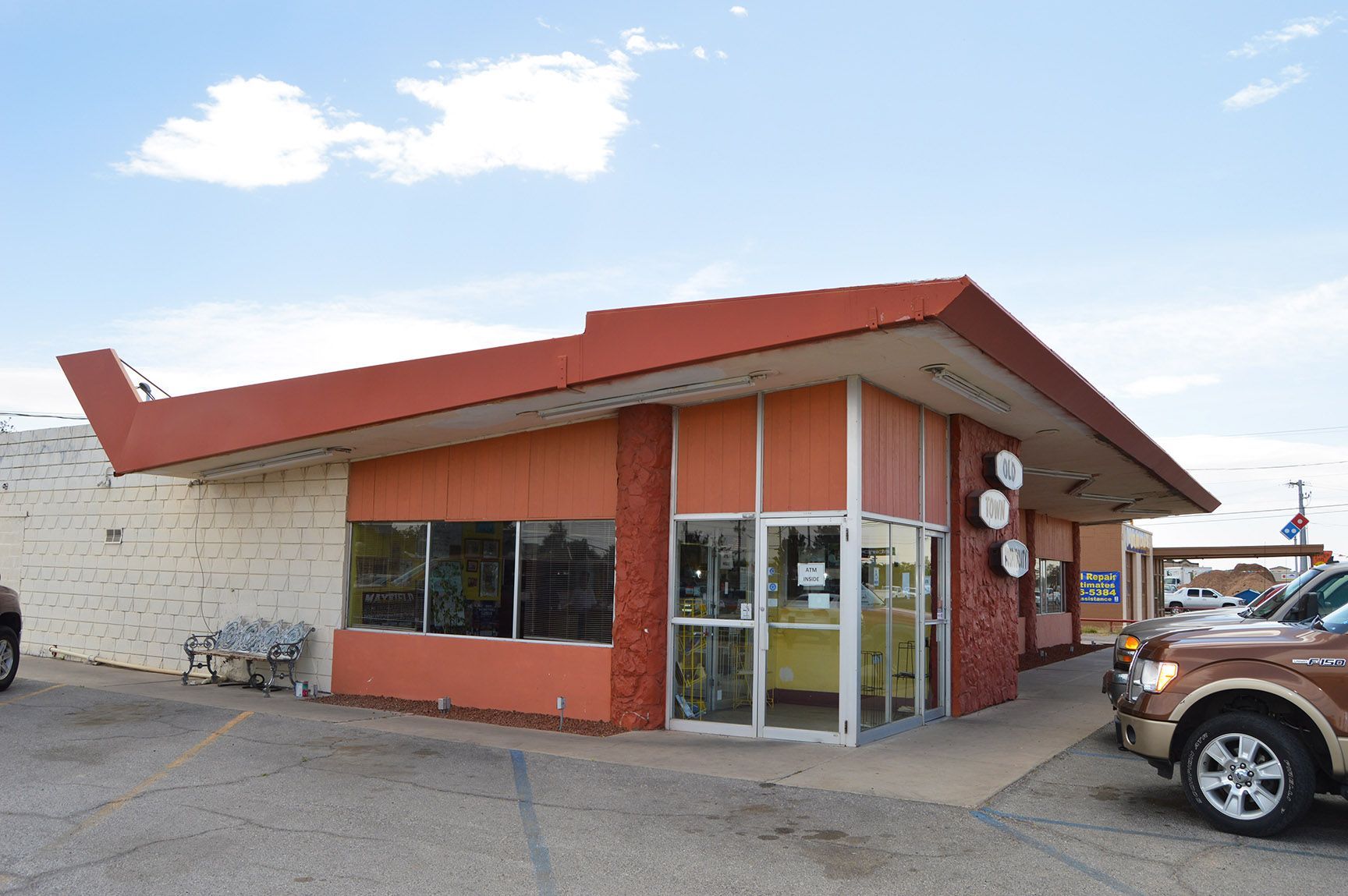 Low red-roofed corner building with glass storefront and parked vehicles under a blue sky