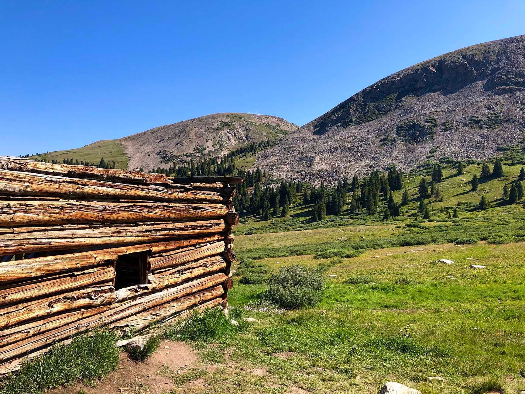 Weathered log cabin beside a green meadow with pine trees and rocky mountains under a clear blue sky, The ruins of an old cabin, Mayflower Gulch, Colorado