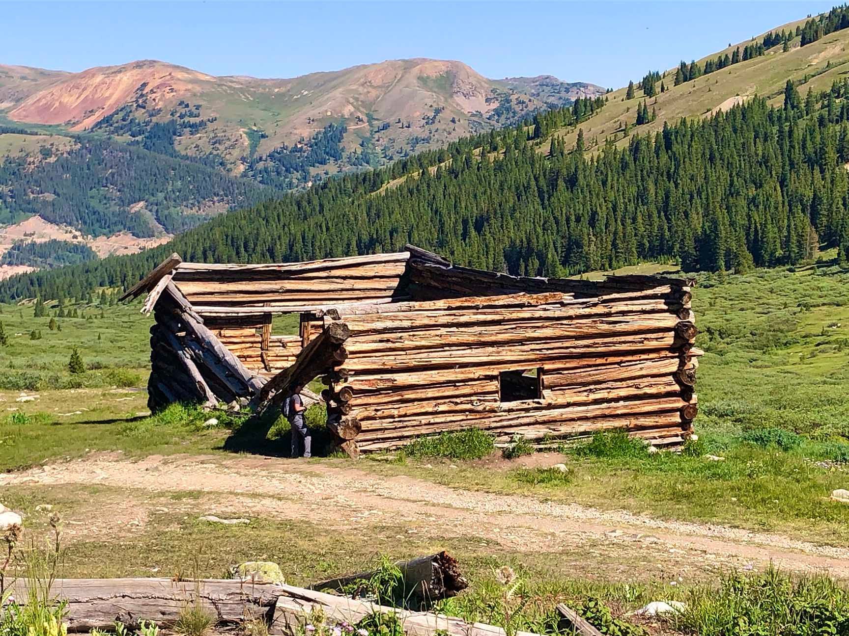Weathered log cabin ruins in a grassy mountain valley with forested hills and rocky peaks behind., Mayflower Gulch, Colorado